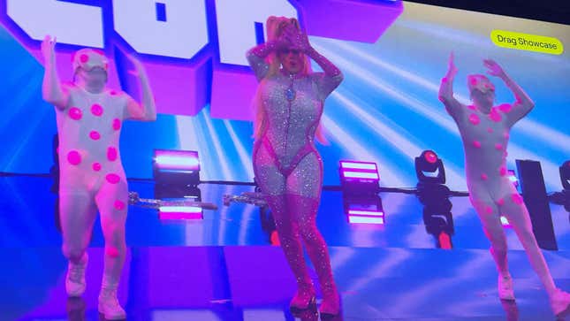 A drag performer dances at TwitchCon while flanked by two dancers in white bodysuits with bright purple spots on them.