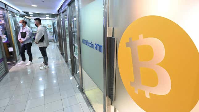A bitcoin symbol on a plane of glass next to HK Bitcoin ATM and two people standing down a hall in the background.