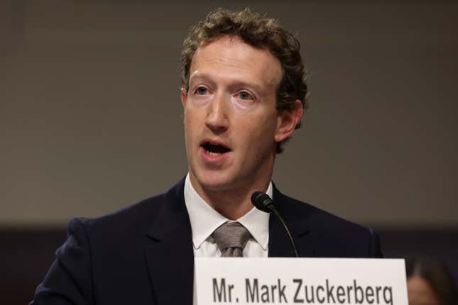 Mark Zuckerberg speaking into a microphone with a Mr. Mark Zuckerberg namecard in front of him
