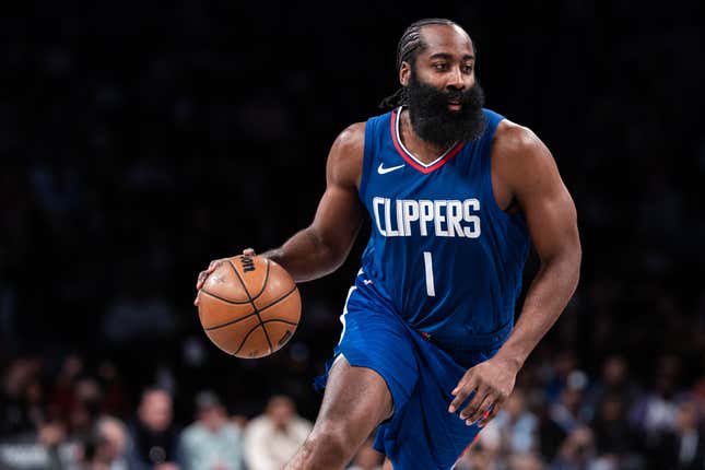 A Black man with with a thick black beard, wearing a blue and white Los Angeles Clippers basketball uniform, dribbles a basketball before a large crowd of fans.