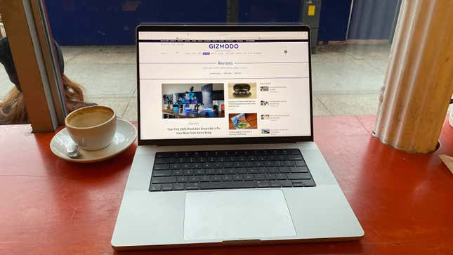 A 14-inch MacBook Pro on a table next to an empty cup of coffee with the main page on screen being Gizmodo.