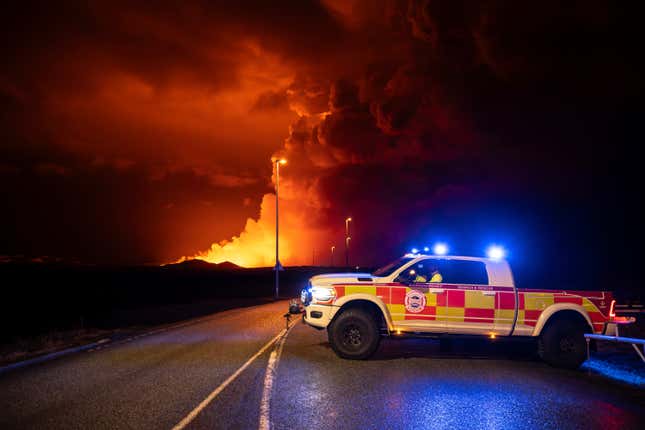 An emergency vehicle sits in the road, with an eruption in background.