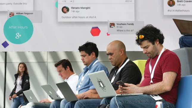 Four conference goers wearing lanyards sit side by side, all with their laptops open