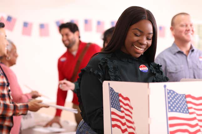 African descent, young or mid-adult woman votes in the USA election. She stands at voting booth in polling station. Other voters and election day registration seen in background.