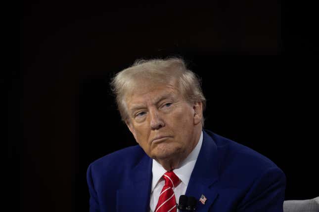 Donald Trump, in a blue suit and red-and-white striped tie, sits in front of a black background