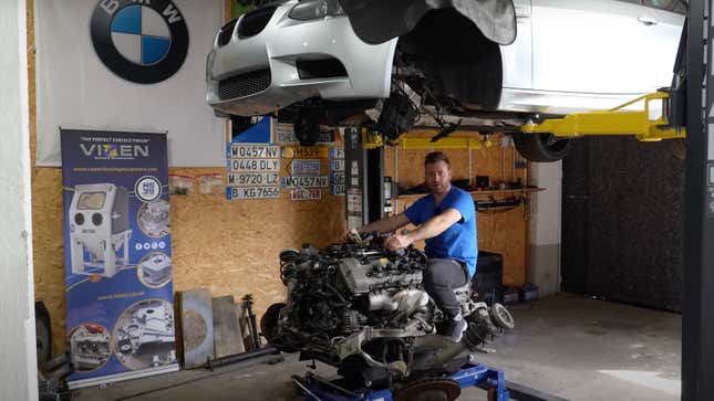 A man in a blue shirt is sitting on an engine and transmission like it's a bull.