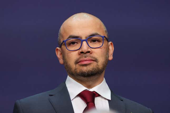 Demis Hassabis wearing blue frame glasses in front of a plain dark purple backdrop