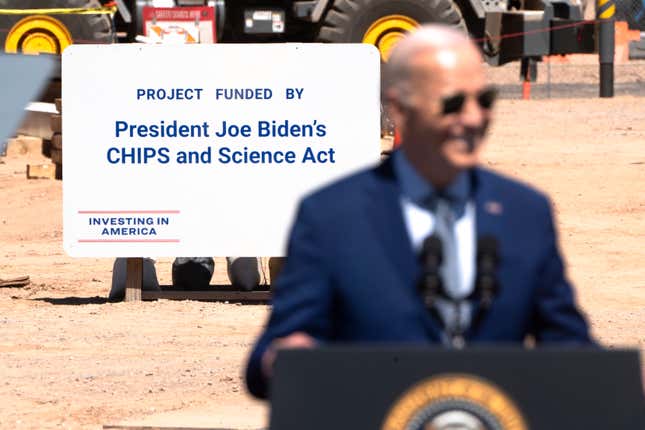Joe Biden is blurry in the foreground standing behind a podium with a partial presidential seal while a sign that says Project Funded By President Joe Biden's CHIPS and Science Act is clear behind him