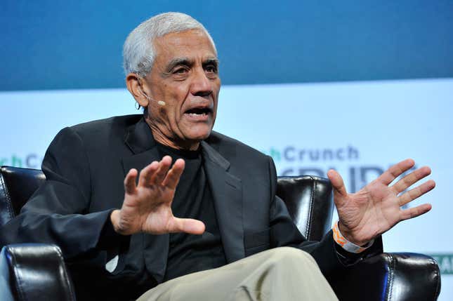 Vinod Khosla wearing a black t shirt and suit jacket with tan pants sitting in a chair and speaking into a lav mic