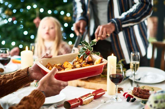 Family Christmas meal with home cooked food being served at dining table, traditional holly with berries garnishing roasted vegetables, Christmas cracker on table with wine and candles.