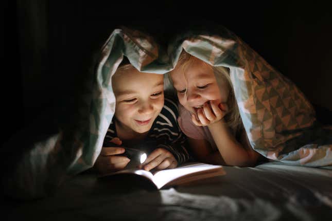 children reading a book with a flashlight under a blanket