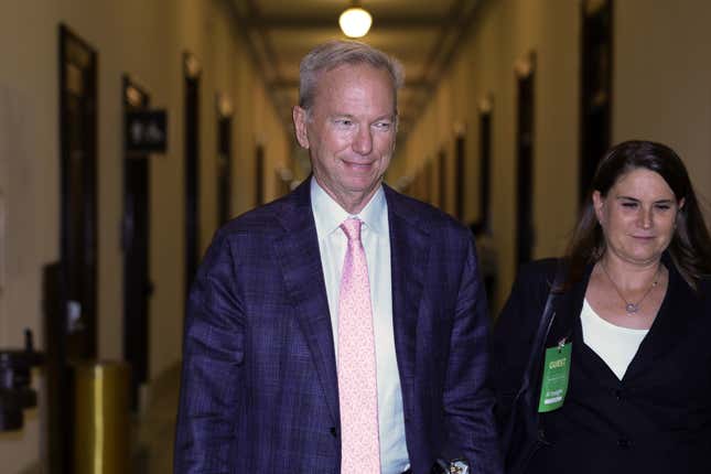 Eric Schmidt wearing a navy suit, white button down, and pink tie walking down a hallway with a brunette woman to his left