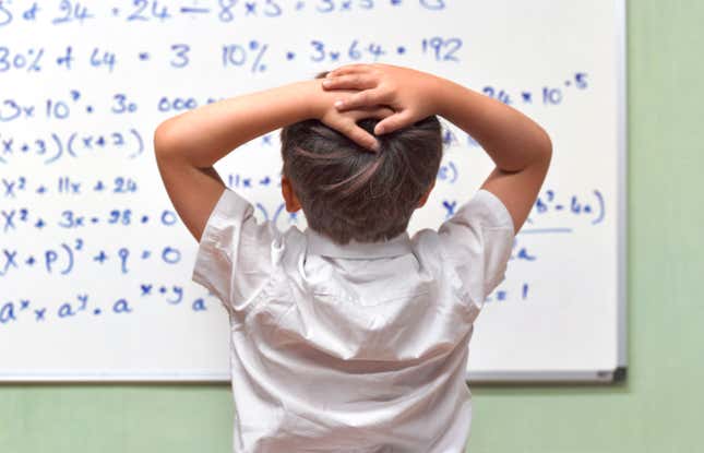 blonde boy holds his head as he looks at a white board with math equations on it