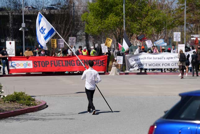 a man holding an Israel flag walking towards protestors with banners calling Google to stop fueling genocide