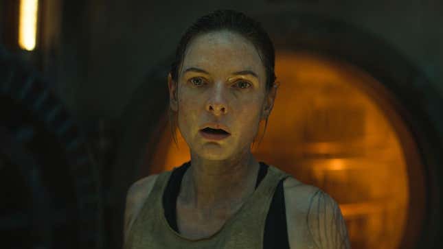 Rebecca Ferguson in Silo, standing in front of a round door to an orange-lit room.