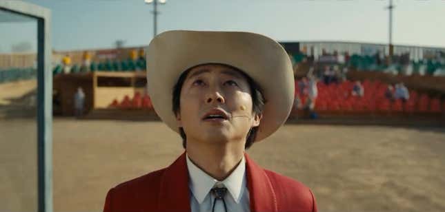 A man (Steven Yeun) wearing a cowboy hat, white shirt with bolo tie, and red jacked looks up at the sky in a scene from Nope.