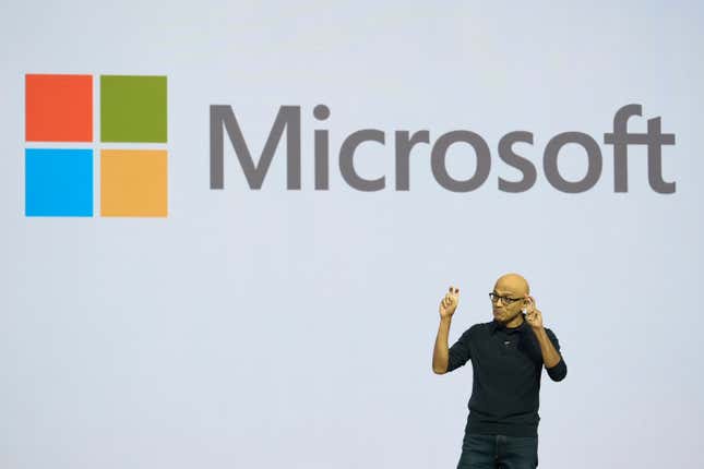 Satya Nadella vistiendo un suéter negro, jeans oscuros, con las manos en alto mientras habla frente a un fondo que dice Microsoft