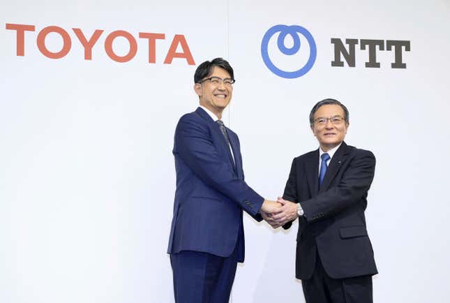 Koji Sato and Akira Shimada both wearing suits, ties, and glasses shaking hands in front of a backdrop with the Toyota and NTT logos