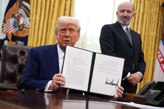 donald trump holding up an executive order with his signature in dark black ink, howard lutnick is standing off to the side behind him looking down at the order