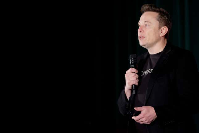 elon musk wearing a black t shirt with an obscured text on it, a black suit jacket, in front of a black background, speaking into a microphone