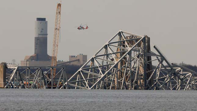 A Coast Guard helicopter flys past a floating crane as it works on the twisted remains of the Francis Scott Key Bridge, which was destroyed when a cargo ship collided with it earlier this week, on March 29, 2024 in Baltimore, Maryland. 