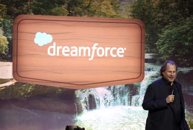 Marc Benioff en la esquina inferior derecha de la foto hablando por un micrófono, de pie frente a un fondo de cascada y un letrero de madera digital que dice dreamforce.