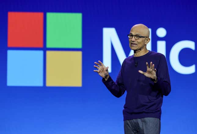 Satya Nadella speaking with his hands up in front of a blue backdrop that has the Microsoft logo