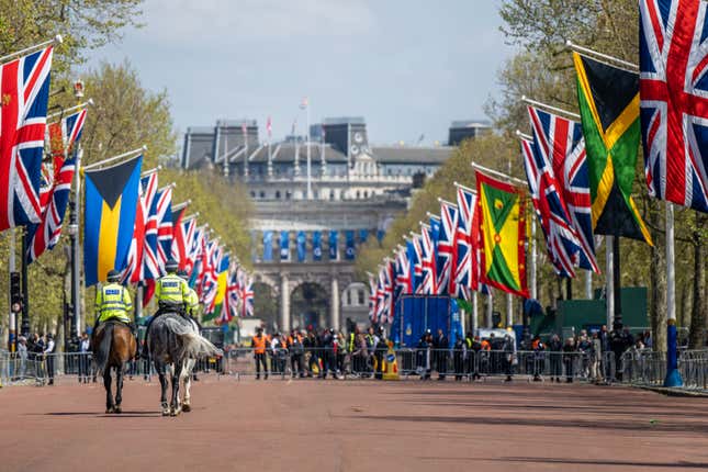 Metropolitan police patrol an intersection near Buckingham Palace on a sunny day. The road is lined with British flags and flags of the Commonwealth countries.