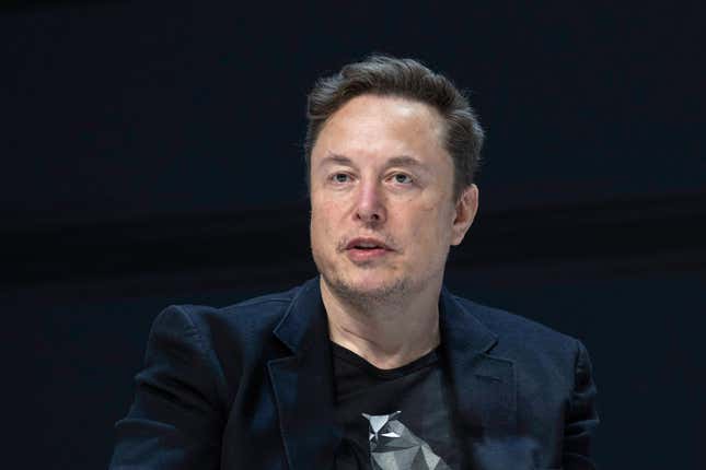 Elon Musk wearing a t shirt and navy suit jacket, speaking