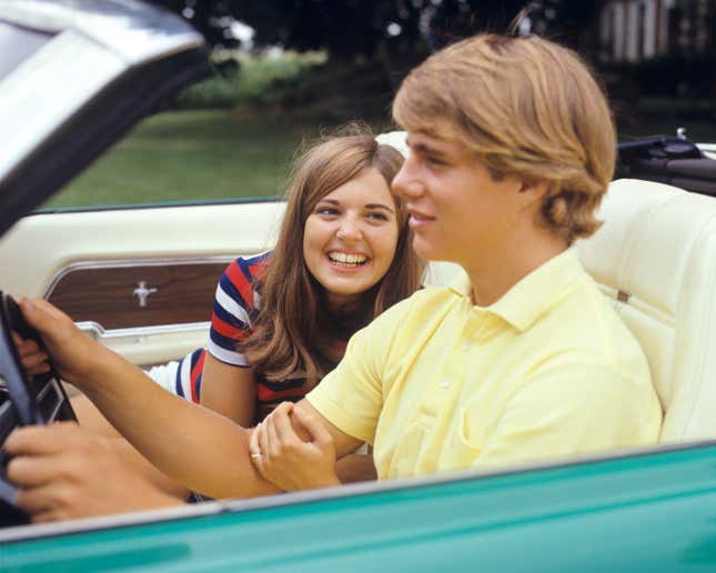 Années 1960-1970 : COUPLE D’ADOLESCENTS SOURIANTS, FILLE FLIRTANT, REGARDANT LE BRAS D’UN GARÇON CONDUISANT UNE VOITURE DÉCAPOTABLE