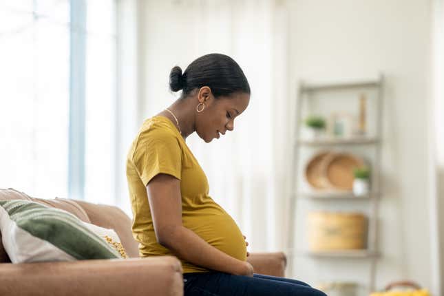 A young pregnant woman of Black woman sits on a sofa in the comfort of her own home as she cradles her belly with her hands. 