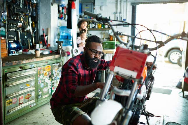 A Black man works on a motorcycle in a garage. A woman and baby look on in the background.