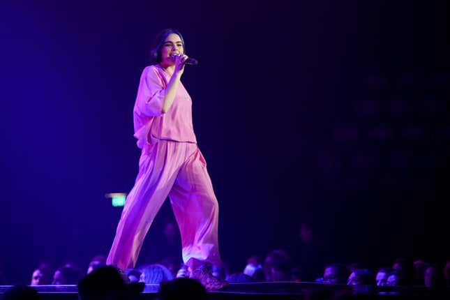 Musician BENEE sings into a mic while walking across stage in a flowy matching pink top and bottom.