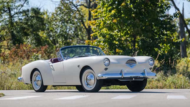 Un Kaiser Darrin blanc avec le toit baissé, garé dans une rue devant des arbres