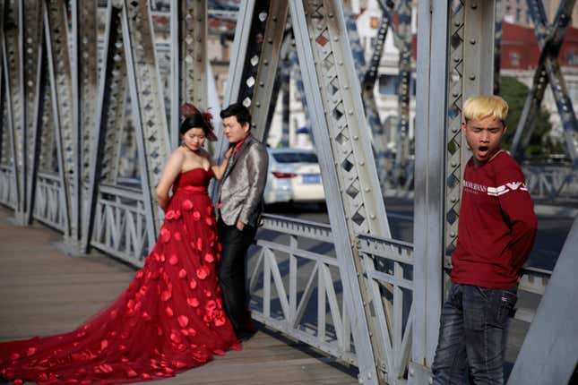 A wedding photographer's assistant in China yawns on the street while couple in formal wear await pictures.