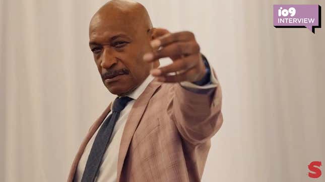 Actor Tony Todd sports a bald head and mustache and is wearing a dark tie and tan suit while gesturing toward the camera in a scene from Shudder's Horror Noire.