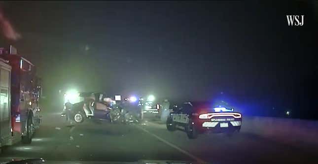 Several police cars and a fire truck sit on the side of a dark freeway. A smashed Tesla Model X sits in the middle of the right hand lane turned across the lane. Emergency lights blur the scene. 