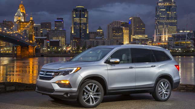 A photo of a silver Honda Pilot crossover with a city skyline in the background. 