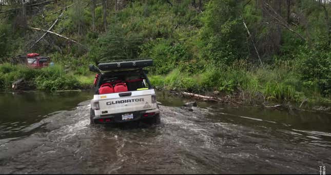 A modified white Jeep Gladiator crosses a river in a dense forest.