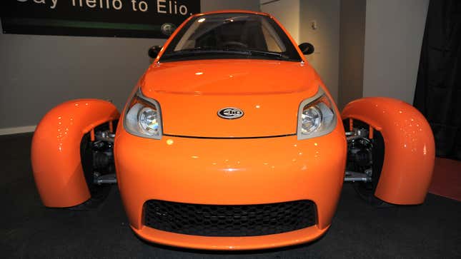 A photo of an orange Elio three-wheeled car. 