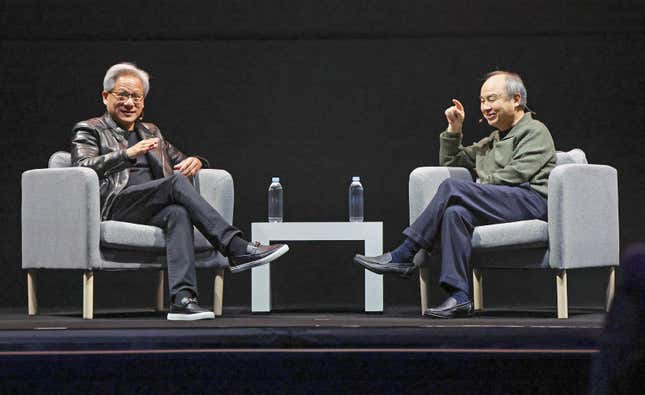 Jensen Huang and Masayoshi Son sitting in grey chairs on a stage with water bottles on a grey table with water bottles