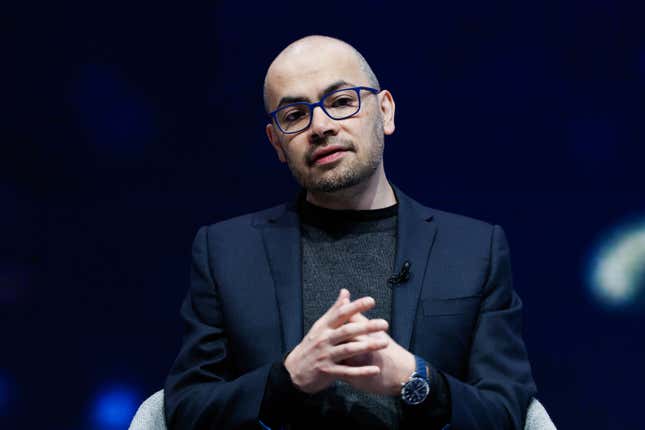 demis hassabis sitting in a chair in front of a dark backdrop with his hands together