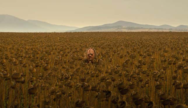 Leatherface's head just peeks above the top of a vast field of tall crops, with mountains in the distance.