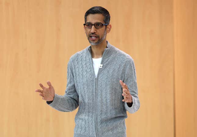 sundar pichai wearing a grey sweater jacket over a white shirt speaking with his hands up in front of a wood wall