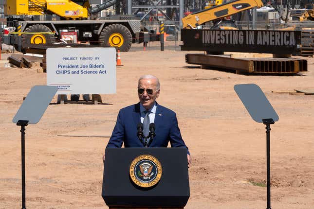 Joe Biden speaking at a podium with the presidential seal, behind him is a bulldozer and a sign about the project funding by the CHIPS and Science Act