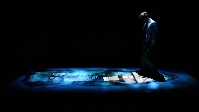 Someone bows their head in a dark room where the layout of the wreck of the HMS Titanic is projected in blue light