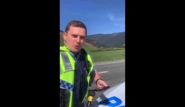 A confused looking New Zealand police officer stands in front of a lush and green landscape