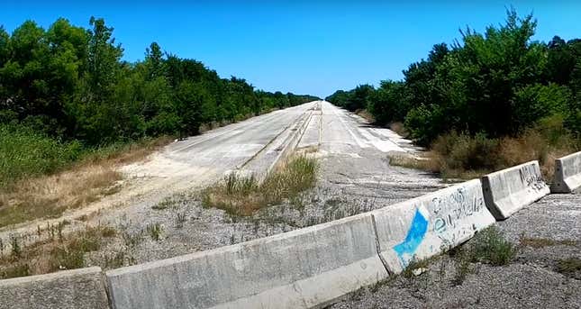 An empty and abandoned road overgrown with weeds stretching off into the distance with barriers blocking vehicle entry