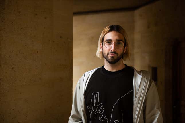 Aidan Gomez lleva gafas y una camiseta negra con un dibujo blanco debajo de una sudadera con capucha grisácea con cremallera y sonríe levemente a la cámara.