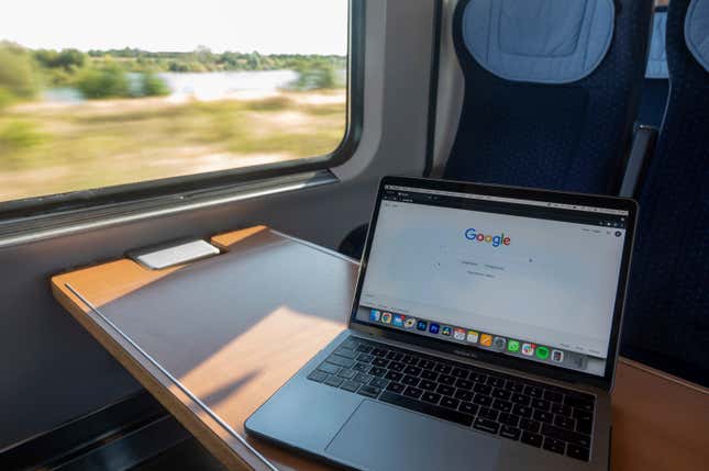 A laptop displays Google's homepage on the table of a traincar rolling through a green german countryside.